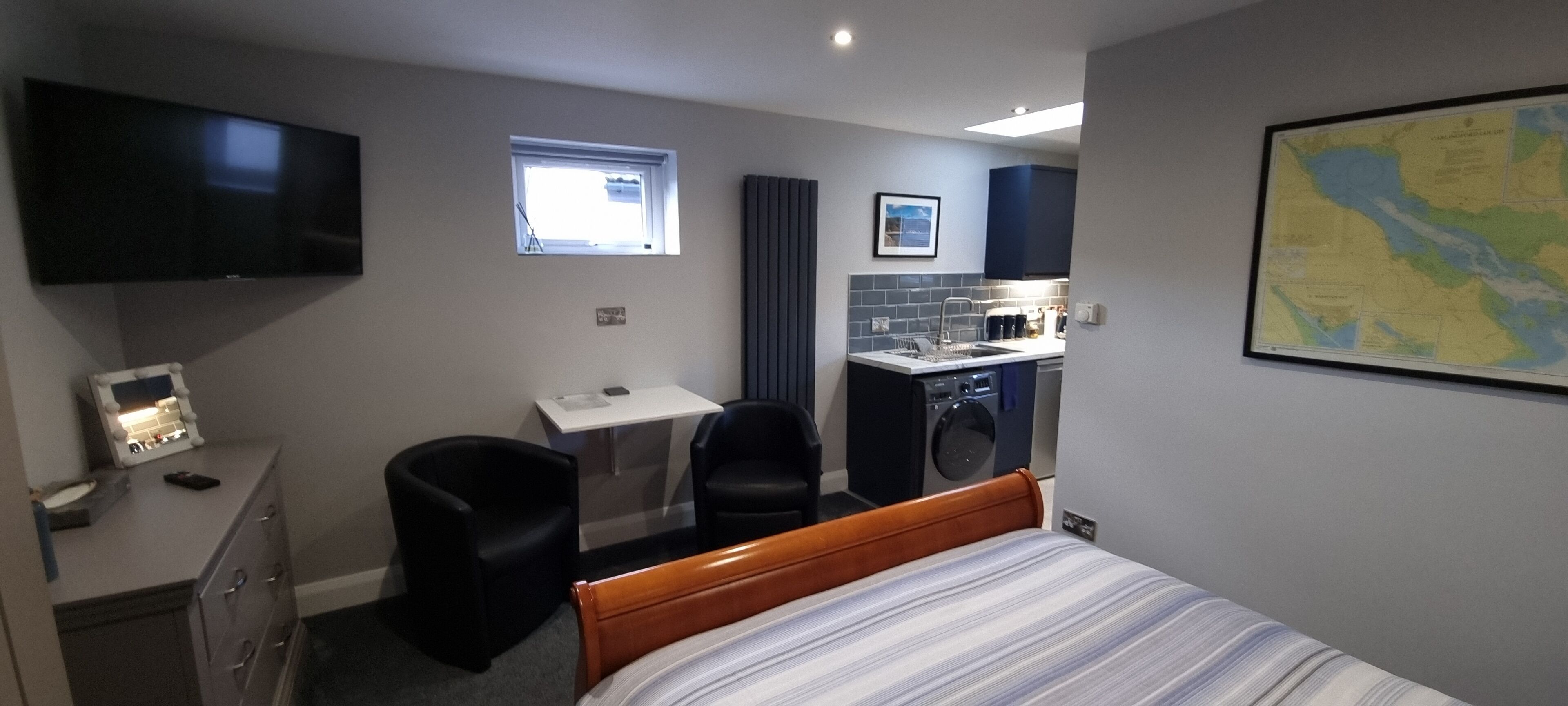 A hotel room with a bed, a desk with a chair, a TV, a small window, a map on the wall, and a kitchenette.