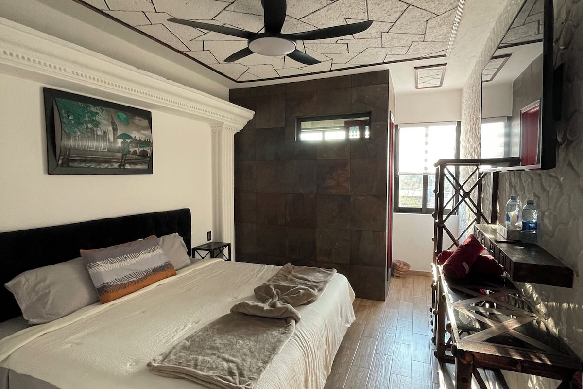 Un dormitorio con cama, ventilador de techo, un cuadro y suelo de madera.