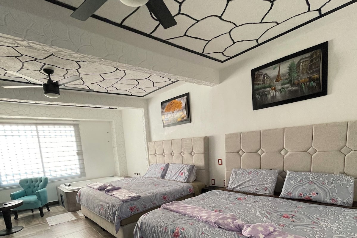 Un dormitorio con dos camas, un ventilador de techo, una ventana con persianas, una silla y un cuadro en la pared.