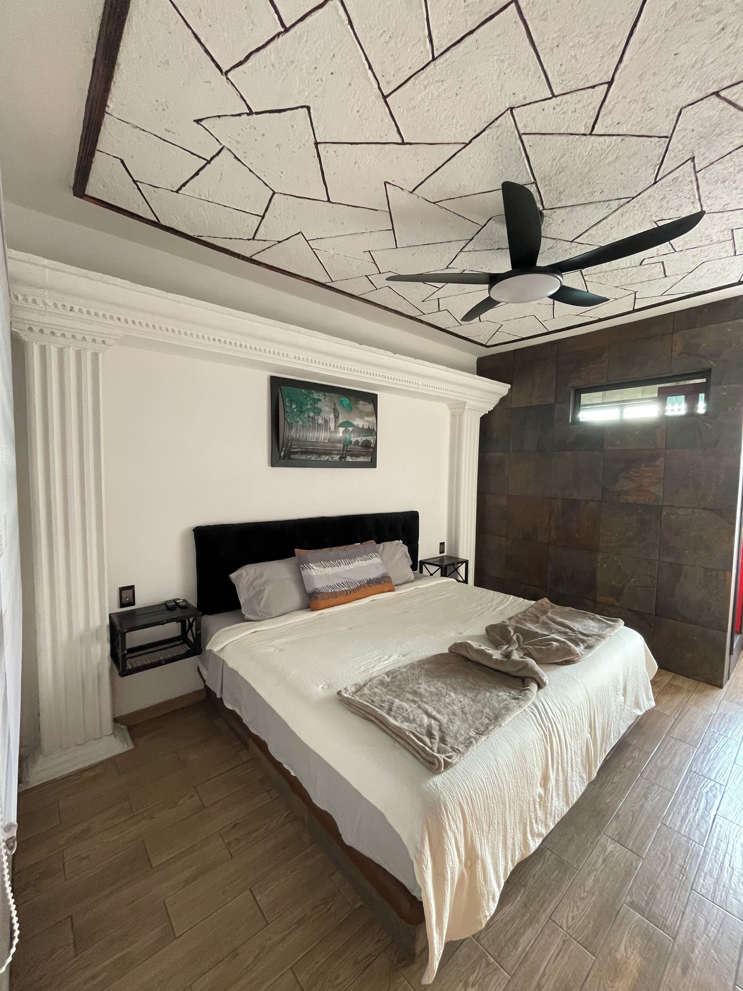 Un dormitorio con una cama grande, un ventilador de techo, una mesita de noche y un cuadro en la pared.