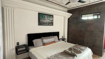 Un dormitorio con una cama grande, un ventilador de techo, una mesita de noche y un cuadro en la pared.