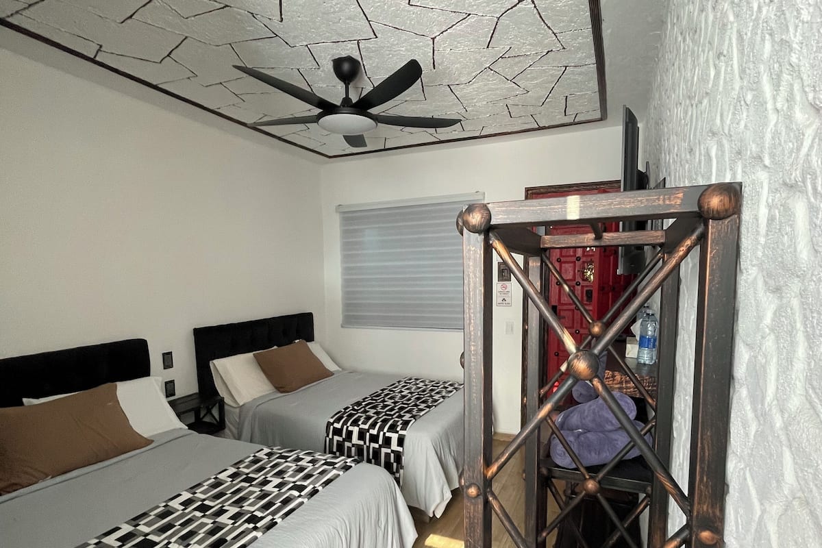 Una habitación con dos camas, un ventilador de techo, una ventana con persianas y una escalera con barandilla de metal.