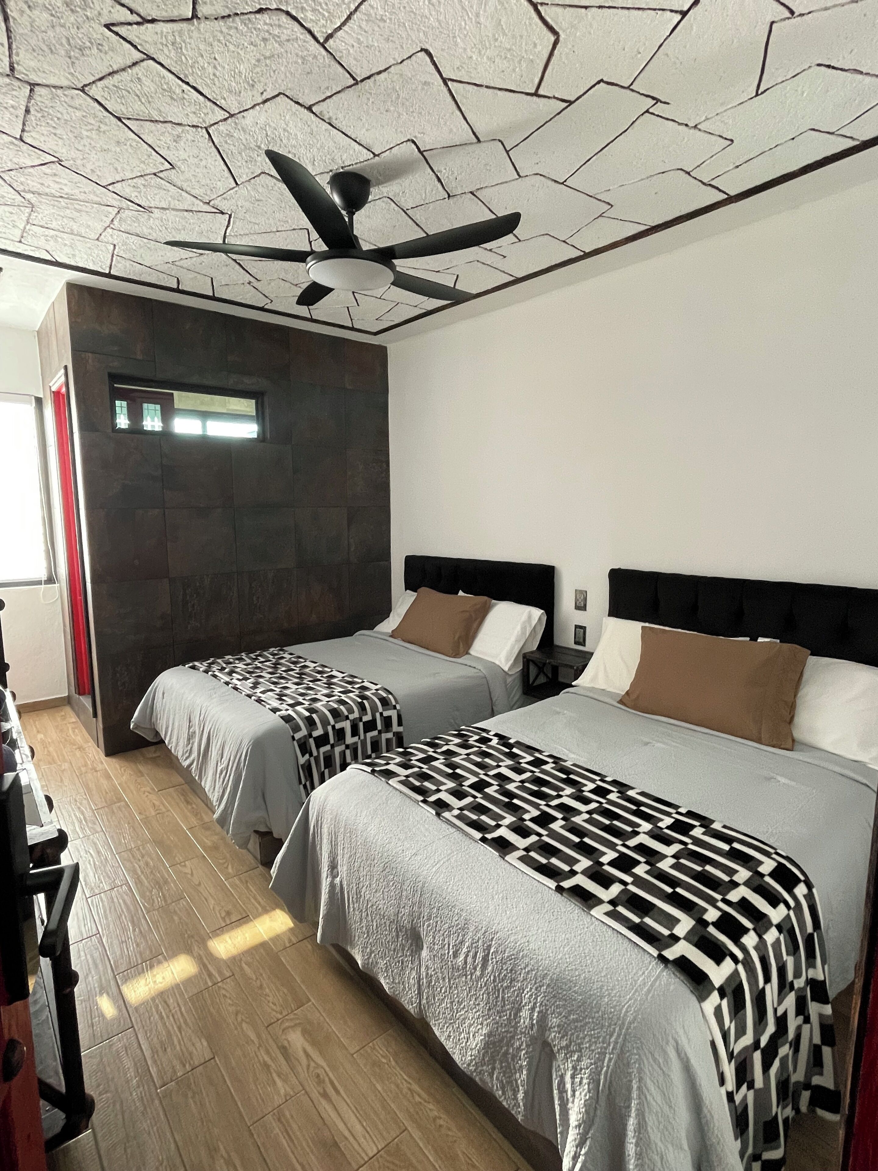 Habitación de hotel con dos camas, ventilador de techo, pared de acento de piedra y ventana con cortinas.