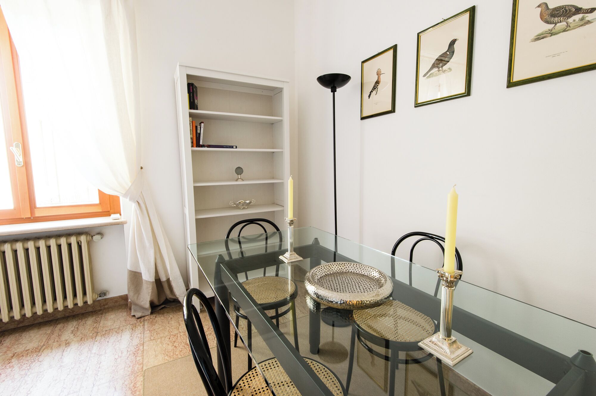 A dining area with a glass table, black chairs, a white radiator, a bookshelf, and framed bird illustrations on the wall.
