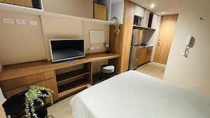 Een moderne hotelkamer met een bed, een bureau met een televisie, een klein krukje, een koelkast en een magnetron.