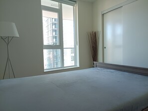 A bedroom with a large window, a bedside table, and a tall vase of dried plants.