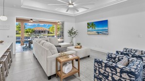 Un salon moderne avec un canapé, une table basse et des fauteuils à motifs. On y trouve un ventilateur de plafond, un tableau représentant une plage, ainsi qu’une vue sur une piscine et un espace extérieur.