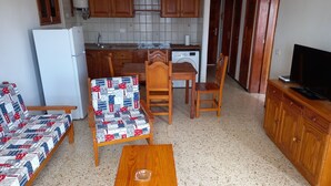 A compact living space with a kitchenette, a sofa with a patterned cover, a wooden dining table, and a television on a wooden cabinet.