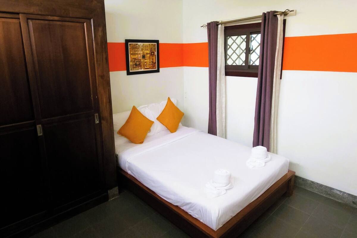 Una cama individual con ropa blanca de cama, dos almohadas naranjas, un cuadro en la pared y una ventana con cortinas moradas.