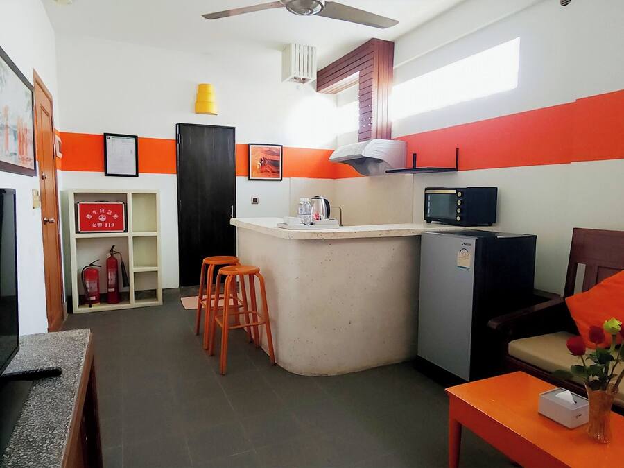 A compact living space with a kitchenette, a sofa with orange cushions, a small table with a vase, and a television.