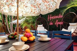Un área de estar al aire libre con una mesa preparada para una comida, rodeada de sillas y un paraguas decorativo.