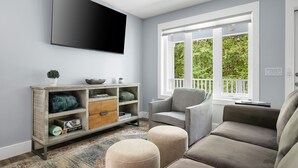 A modern living room with a flat-screen TV, a grey sofa, ottomans, a wooden console, and a large window with a view of greenery. - Private & Quiet Retreat Near Folly Beach (Charleston)
