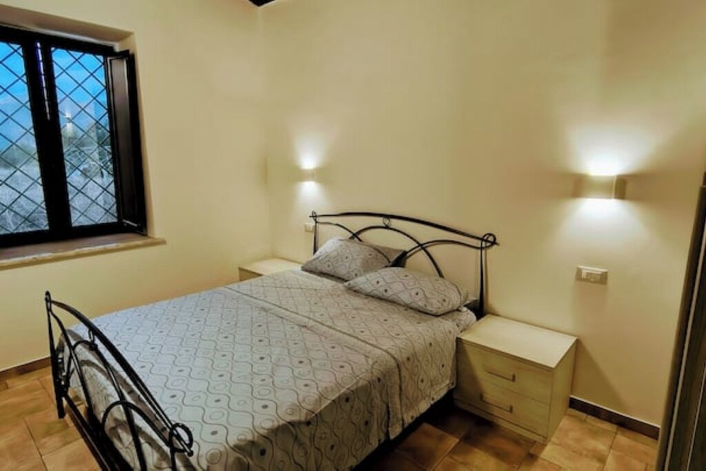 A single bed with a patterned bedspread and pillows, a bedside table, a nightstand, a window with a metal grille, and a wall-mounted light fixture.