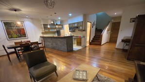 A spacious living area with a wooden floor, a sofa, a coffee table, and a dining table with chairs. There is a kitchen area with a counter and cabinets, and a staircase leading to another level. - Beautiful apartment with incredible view. Super spacious, comfortable and full of light (Cochabamba)