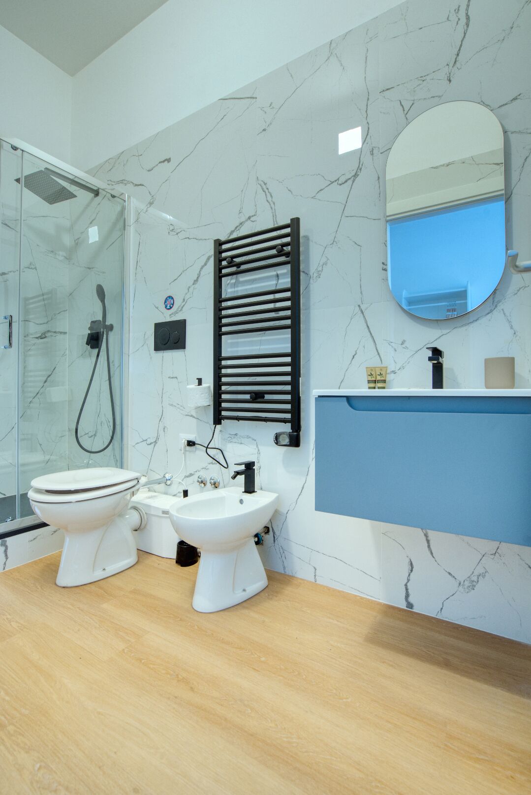Et moderne badeværelse med brusekabine i glas, to toiletter, et bidet, en vask og et spejl.