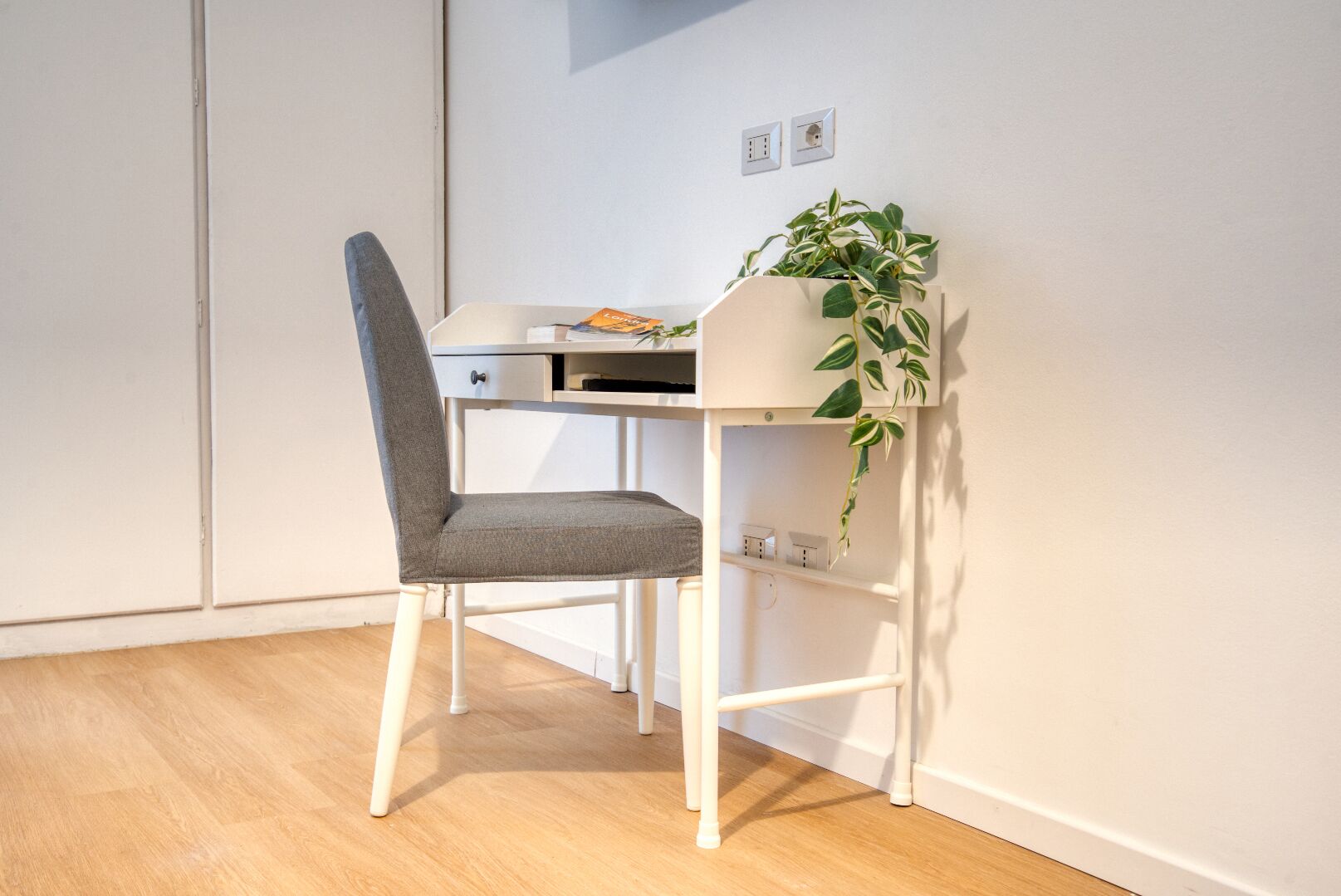 A small, white desk with a grey chair, a potted plant, and a white wardrobe in the background.