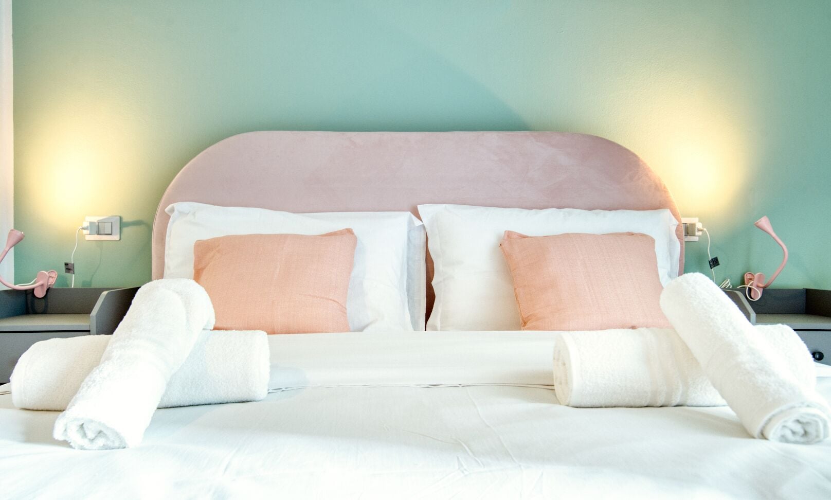Ein ordentlich bezogenes Bett mit weißen Laken und zwei pfirsichfarbenen Kissen, ein rosa Kopfteil sowie zwei Nachttischlampen.
