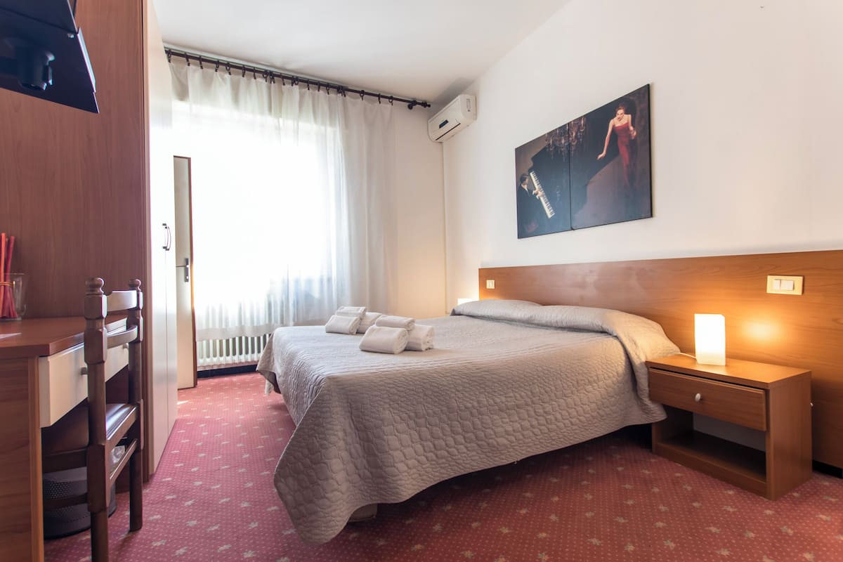 A hotel room with a bed, a nightstand with a lamp, a wooden desk, a chair, a radiator, and a window with curtains.