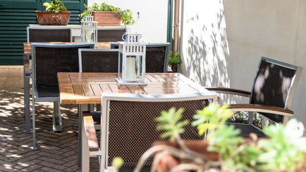 Un patio al aire libre con una mesa de madera y sillas de metal, rodeado de plantas en macetas y un farol blanco.