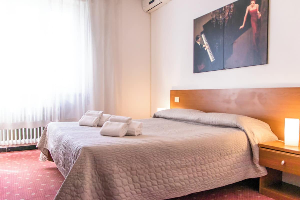 A hotel room with a large bed, a bedside table, a nightstand, a wall-mounted artwork, and a window with curtains.