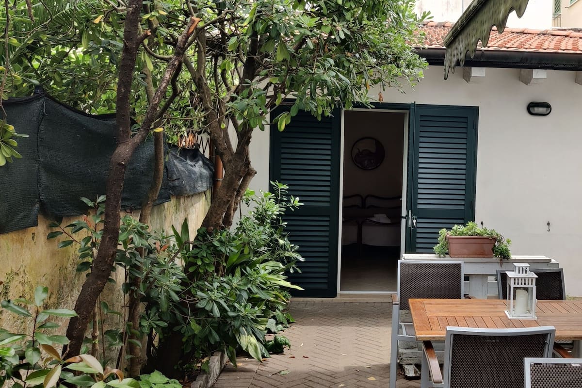 An outdoor patio with a wooden table and chairs, surrounded by greenery and a building with green shutters.