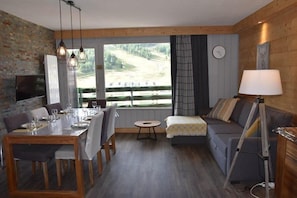 Quarto aconchegante com mesa de jantar posta para quatro pessoas, um sofá, um abajur e uma janela grande com vista para uma área arborizada.