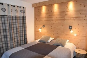 Quarto com cabeceira de madeira, duas luminárias de apoio, cama com cortina xadrez e uma pequena mesa redonda.