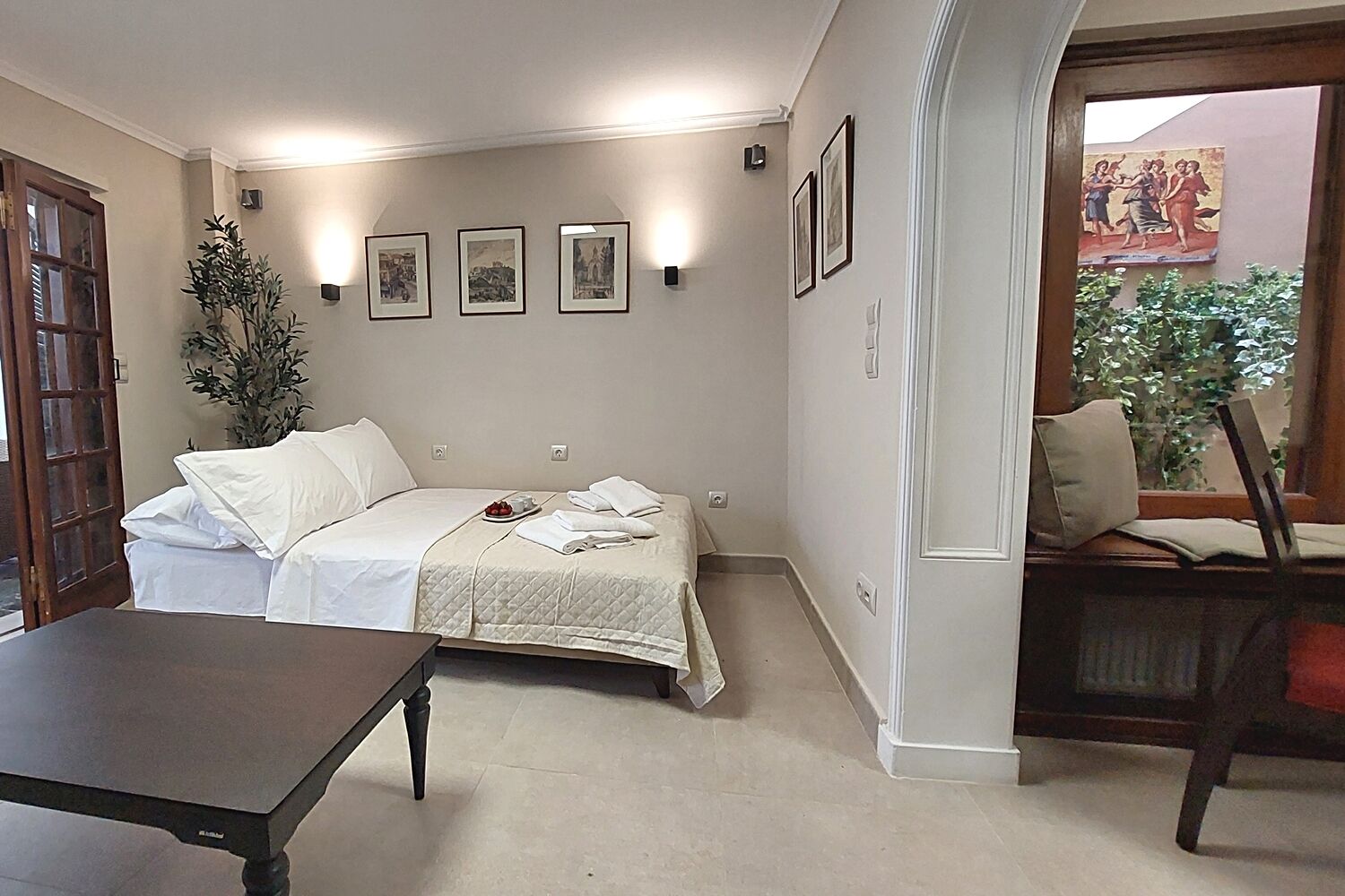 A hotel room with a bed, a desk, and a chair. There are framed pictures on the wall and a window with a view of greenery.