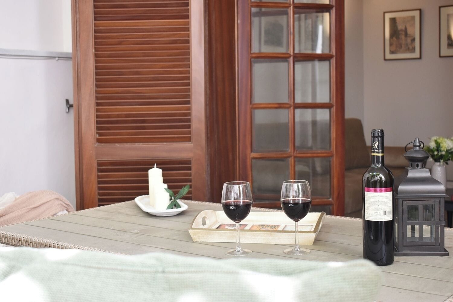 Une table en bois, avec deux verres à vin et une bouteille de vin, une bougie et une lanterne, posés sur une véranda.