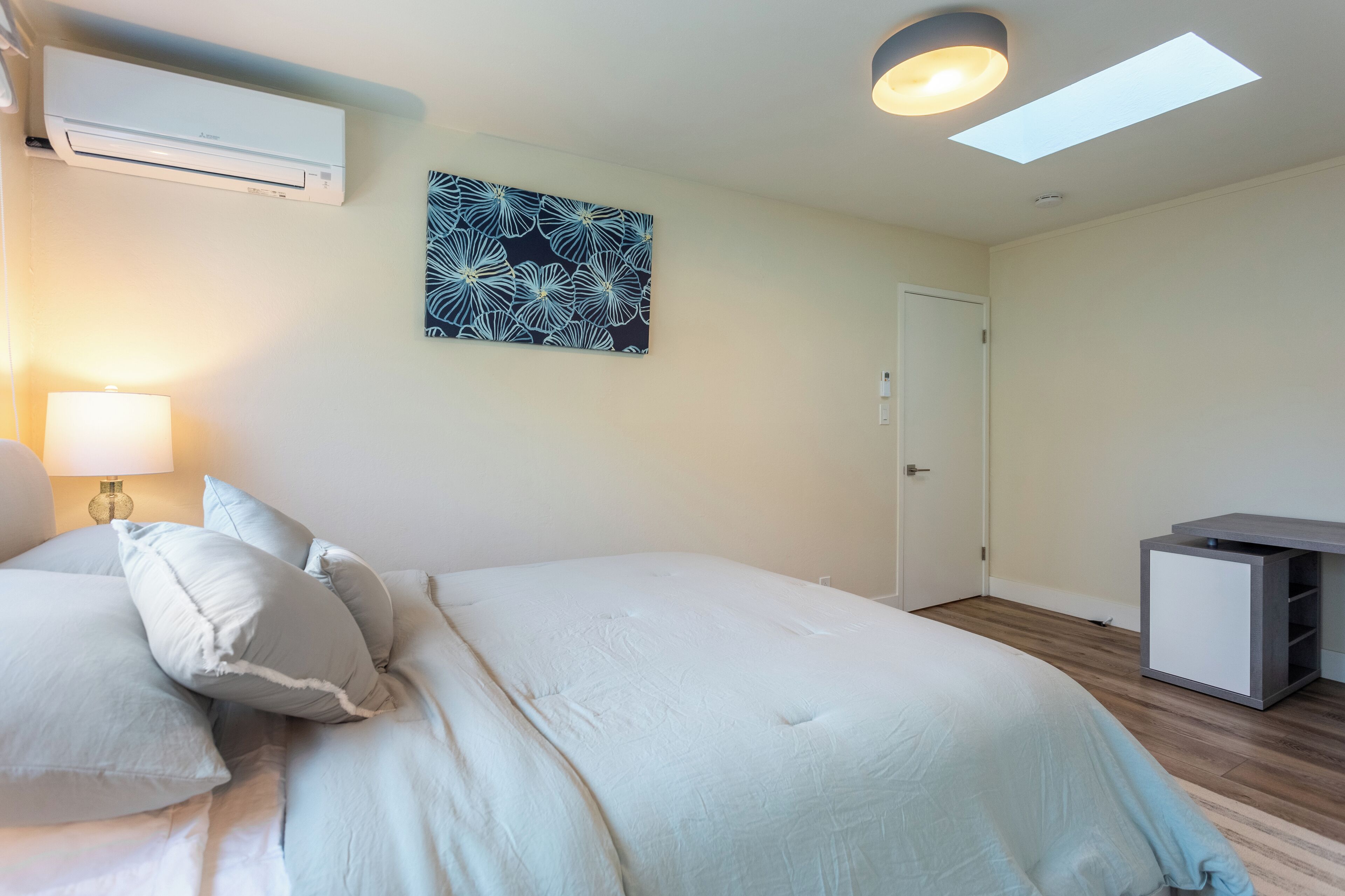 A bedroom with a bed, pillows, a bedside lamp, an air conditioning unit, a door, and a small desk with storage.