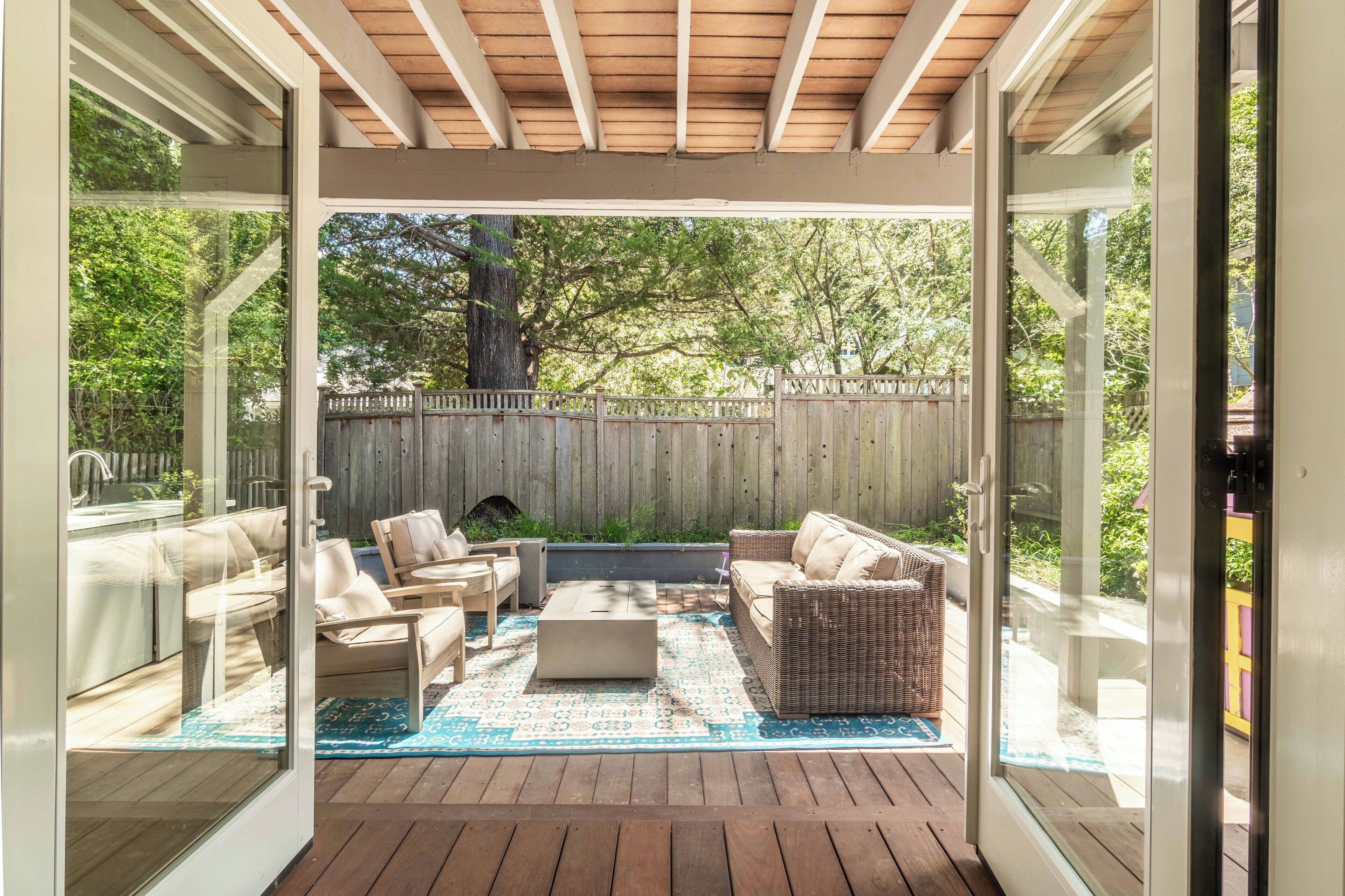 Une terrasse extérieure dotée d'une plateforme en bois, de meubles en osier et d'une porte vitrée donnant sur un jardin.
