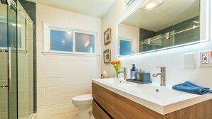 Ein modernes Badezimmer mit Duschkabine aus Glas, Toilette in Weiß, Holzwaschtisch mit weißem Waschbecken und einem großen Spiegel.