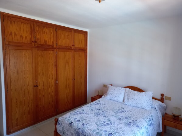 A bedroom with a large wooden wardrobe, a bed with floral bedding, and a nightstand with a lamp.
