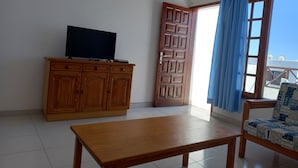 Una habitación con un armario de madera, un televisor encima, una mesa de madera y una ventana con cortina azul.