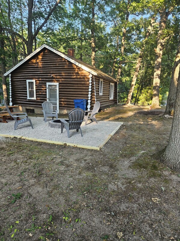 A rustic log cabin with a gravel patio, two Adirondack chairs, and a blue trash bin.