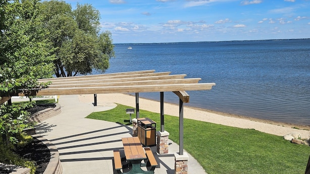 Une terrasse au bord de l’eau, avec des tables de pique-nique et une pergola offrant une vue sur un lac.