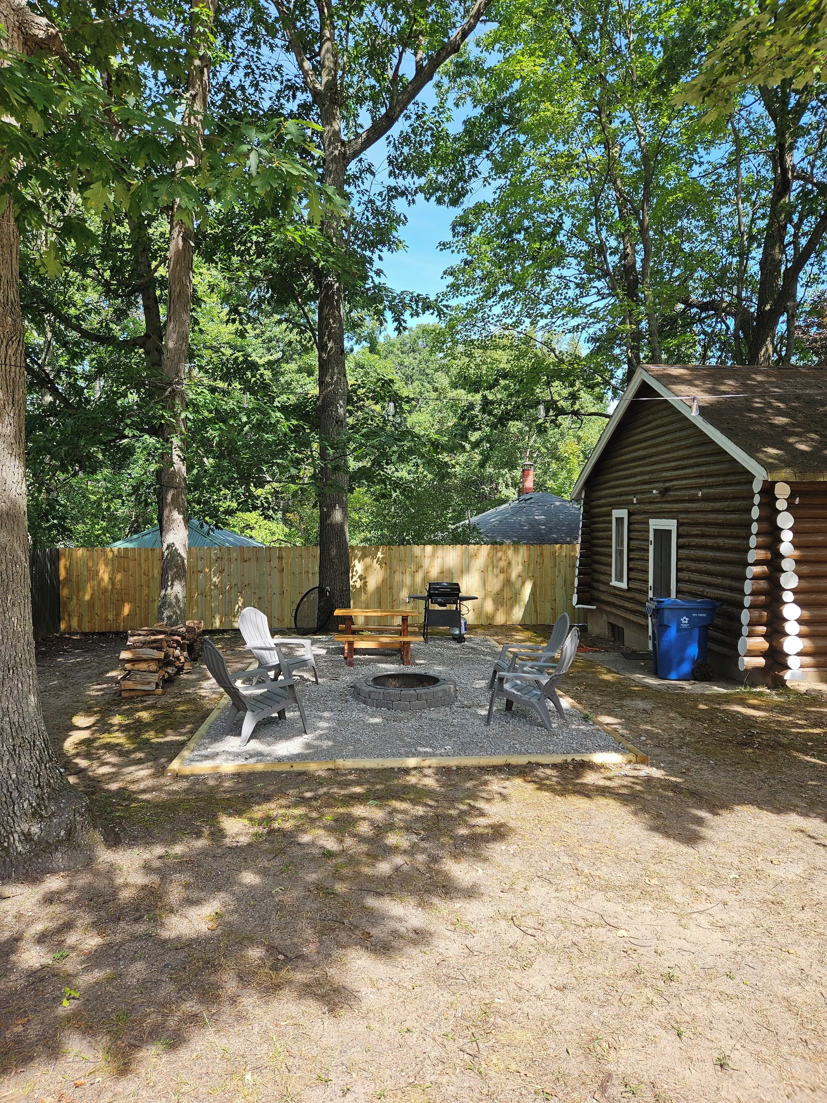 A rustic wooden cabin with a fire pit, picnic table, and Adirondack chairs in a wooded backyard.