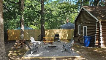 A rustic wooden cabin with a fire pit, picnic table, and Adirondack chairs in a wooded backyard.