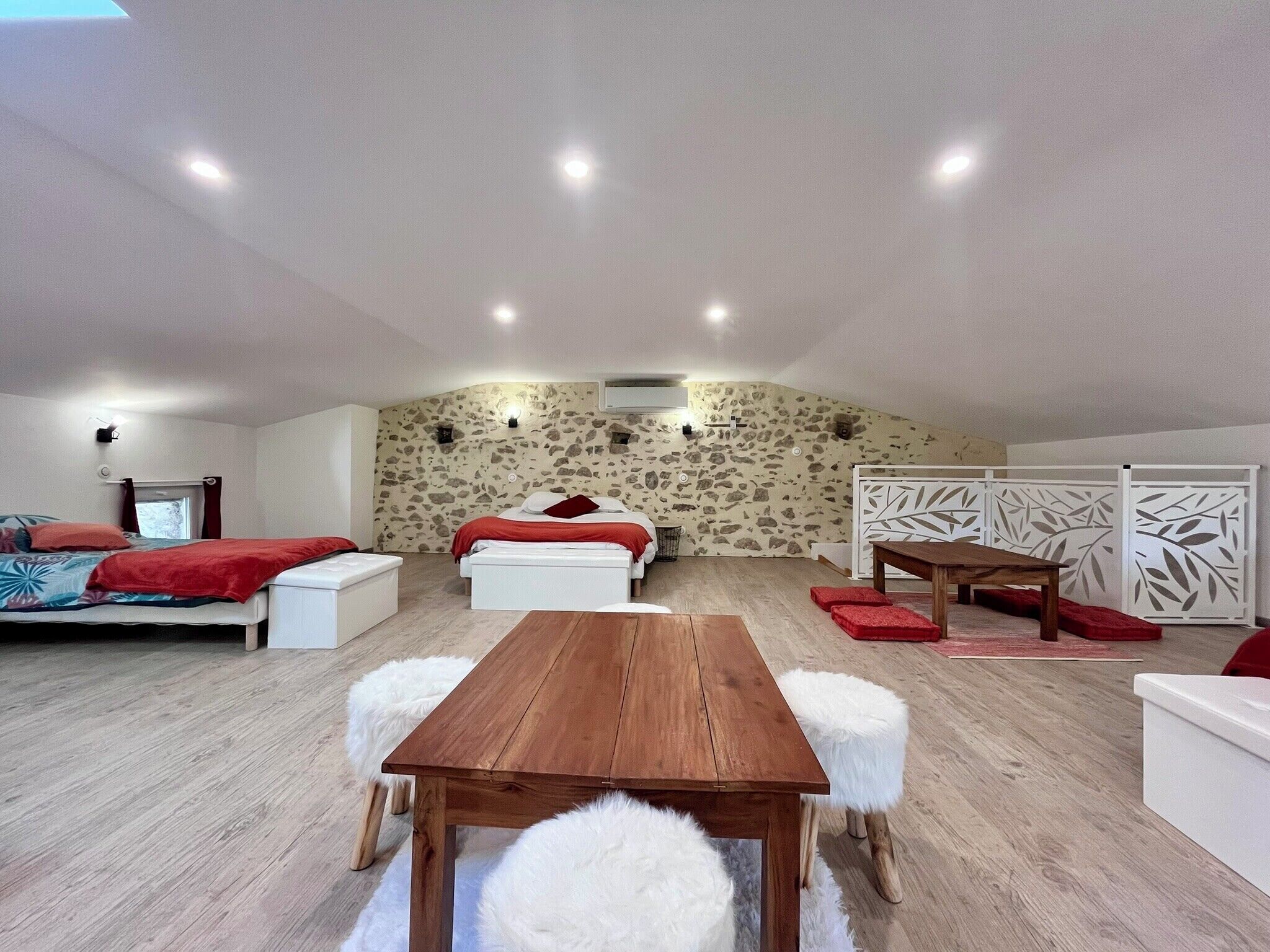 Quarto aconchegante e bem iluminado com mesa de jantar de madeira, banquinhos brancos e fofos, um sofá e uma cama com roupa de cama vermelha.