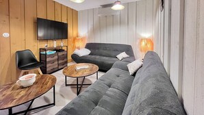 Un salon moderne avec un canapé gris, des murs recouverts de panneaux en bois, un téléviseur à écran plat et des luminaires suspendus.