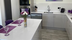 Una cocina moderna con armarios blancos, horno integrado y lavavajillas. Hay una zona de comedor con una mesa blanca y manteles individuales morados, y un jarrón con flores sobre la mesa.