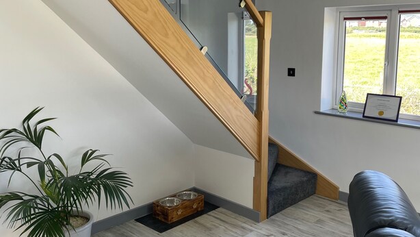 Un interior moderno con una escalera de madera, una planta en maceta, un rincón para la alimentación de mascotas y vistas al exterior a través de una ventana.