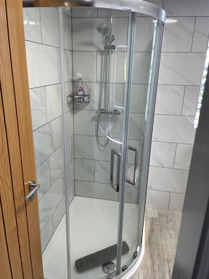 Une cabine de douche fermée par des parois en verre, équipée d’un pommeau de douche à main, d’un flexible de douche et d’un distributeur de savon placé sur une étagère.