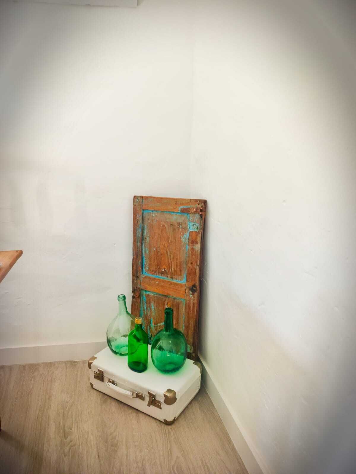 Rincón con una maleta blanca y tres botellas de vidrio verde sobre una superficie de madera.