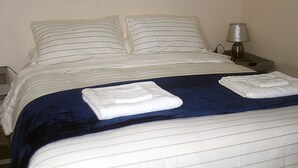 Een keurig opgemaakt bed met een blauwe deken en twee kussens, een bedlampje en een stopcontact op het nachtkastje.