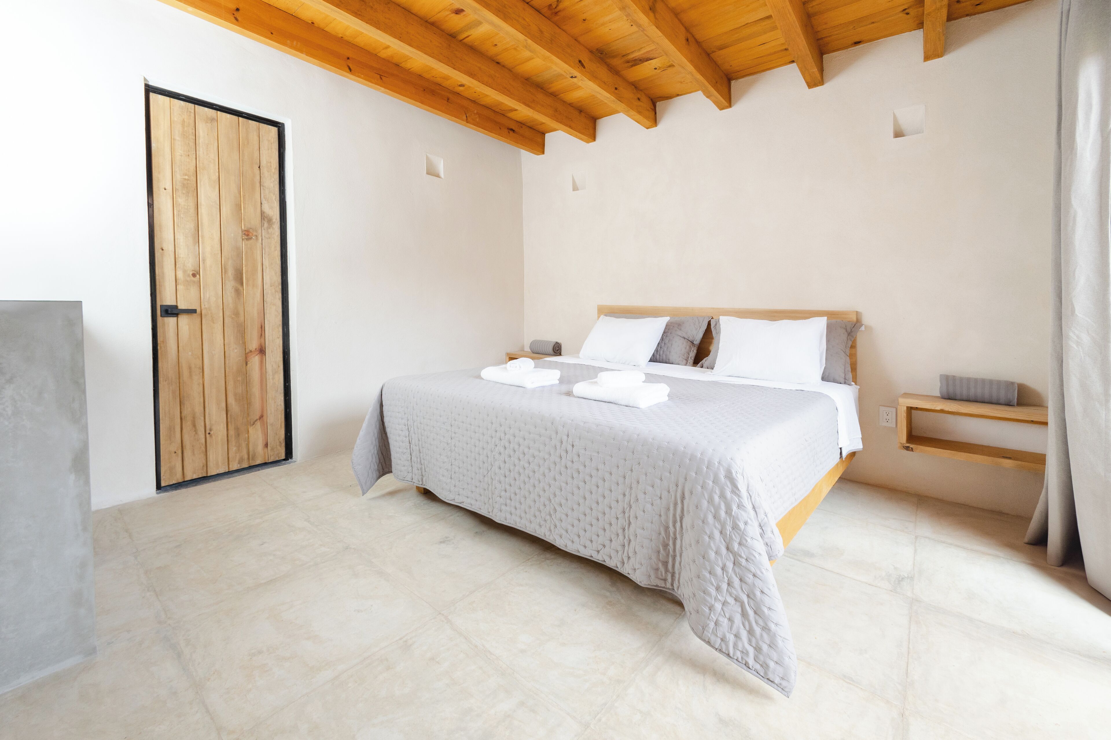 Un dormitorio con techo de madera, cama con ropa de cama blanca, puerta de madera y un estante pequeño con una toalla.