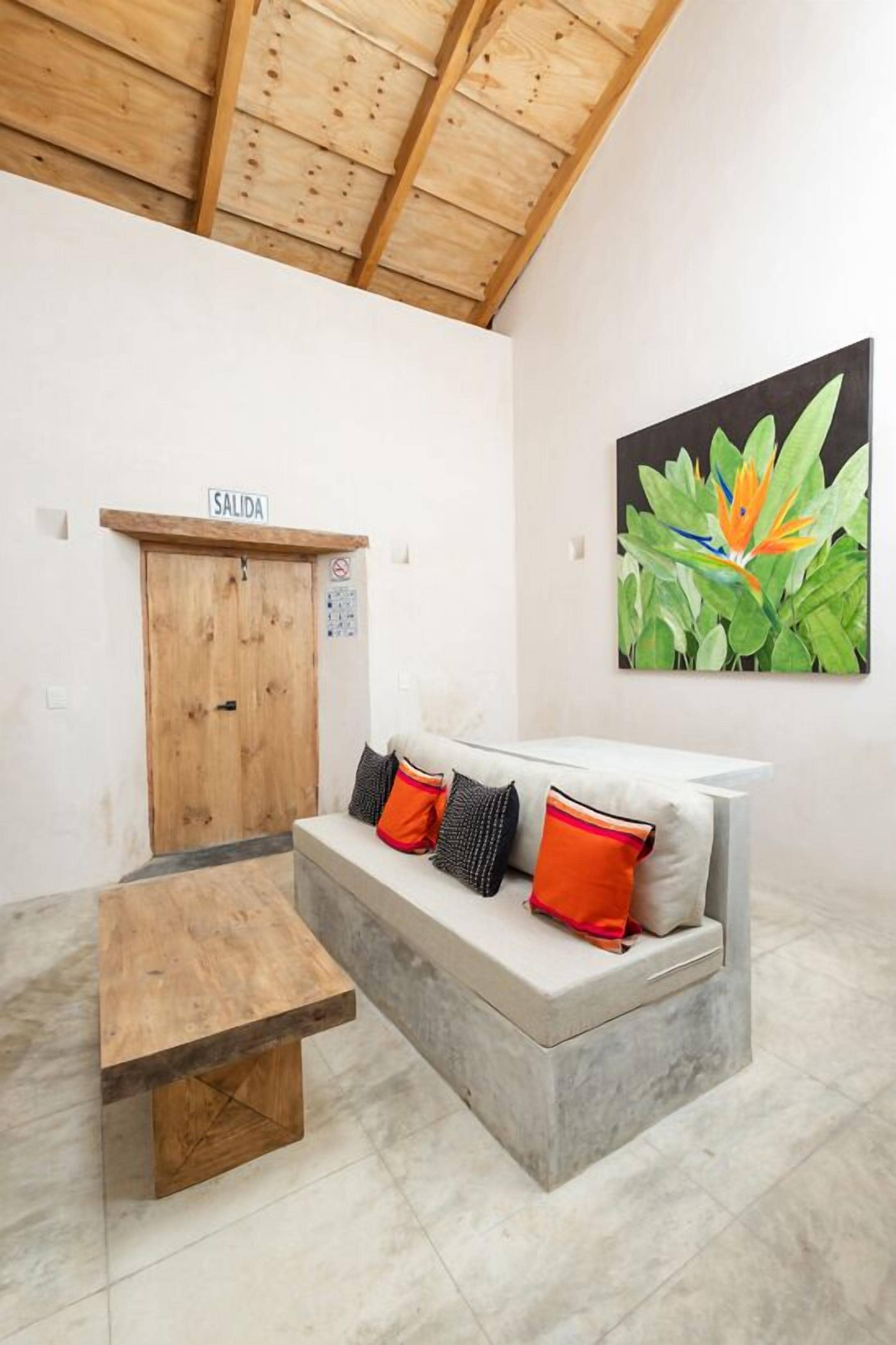 A room with a wooden ceiling, a concrete bench, a sofa with cushions, a door labeled 'SALIDA', and a large flower painting on the wall.