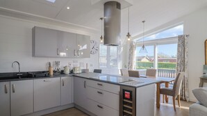 A modern kitchen with a central island, a dining area with a view, and a large window. - Cedar 1 (Axminster)
