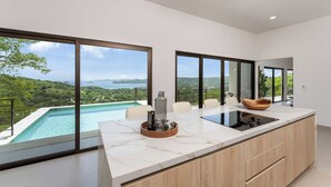 A modern kitchen with a large island, a built-in stove, and a view of a pool and mountains. - Stunning ocean view house in CocoBay Estates (Coco)