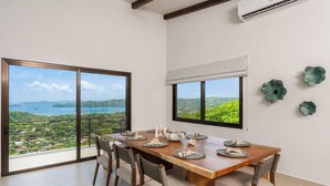 A dining area with a wooden table set for six, large windows with views of greenery and water, and decorative wall art. - Stunning ocean view house in CocoBay Estates (Coco)